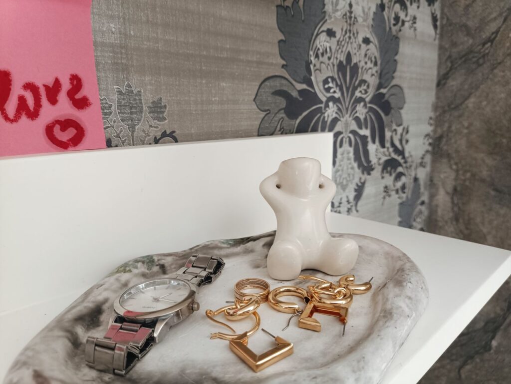A stylish display of jewelry and watch with a ceramic figurine on a marble tray against intricate wallpaper.